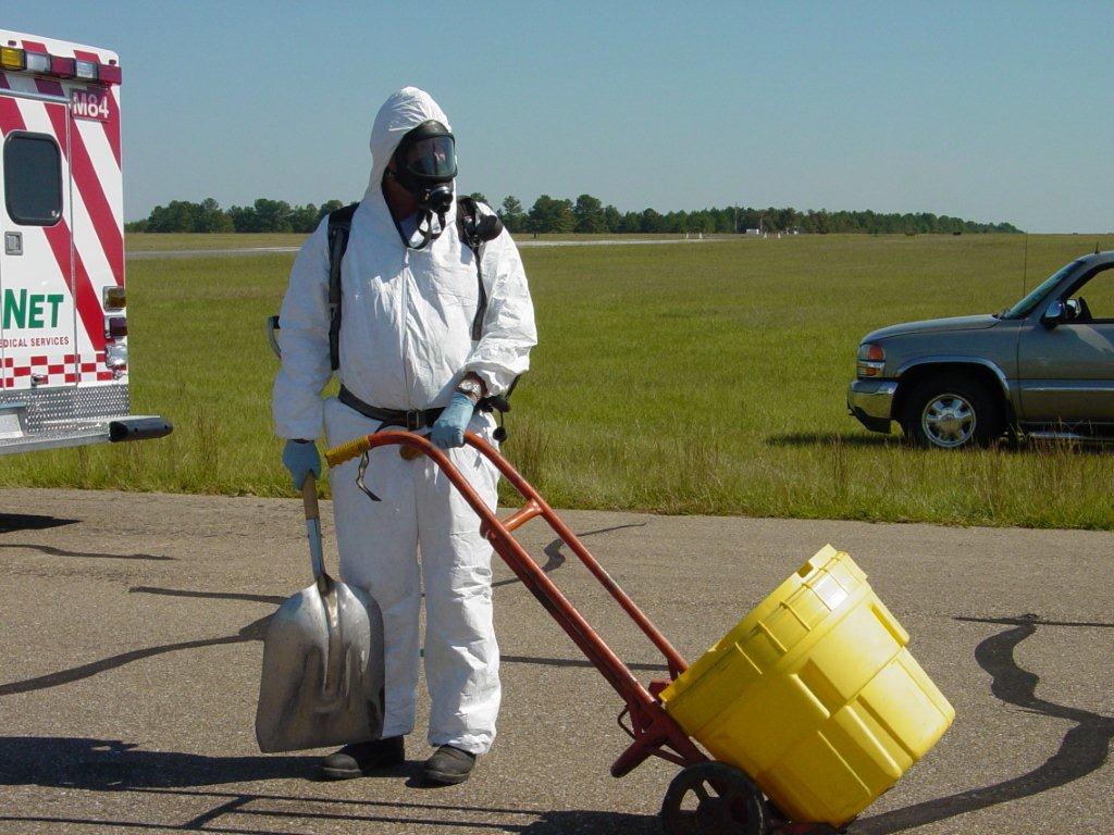 An officer in a hazmat suit, pulling a container for dangerous materials on a dolly.